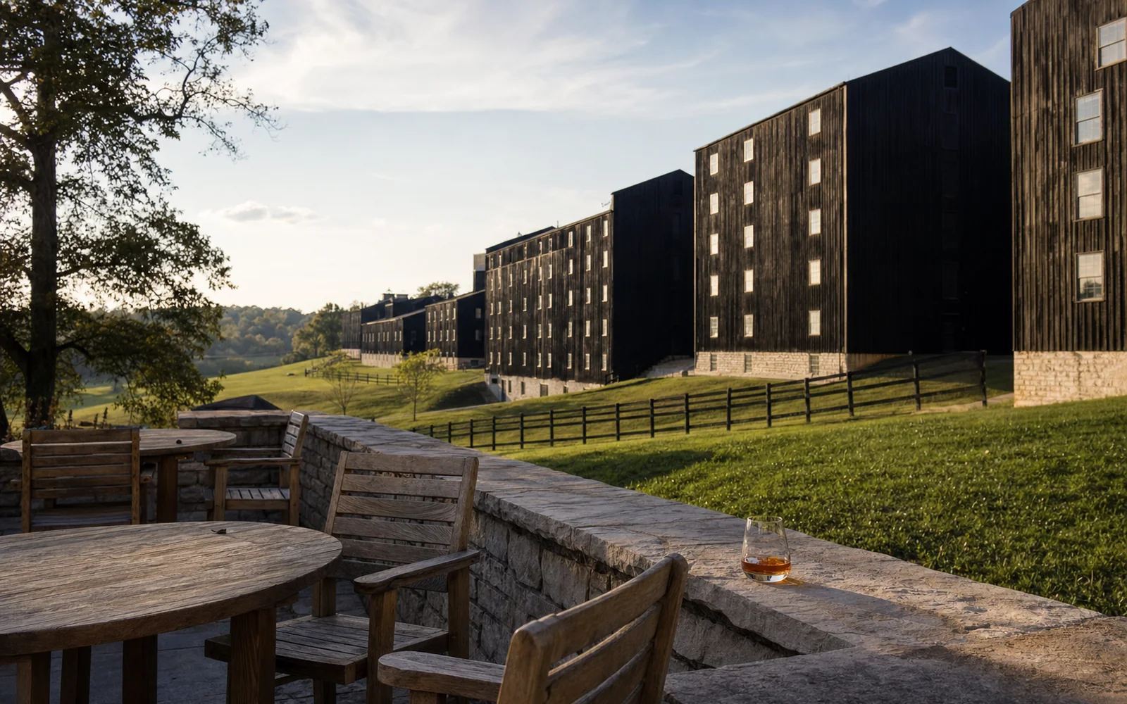 Rows of dark-sided bourbon rickhouses on a green Kentucky hillside under a late-afternoon sky, seen from a stone patio, AI-generated illustration