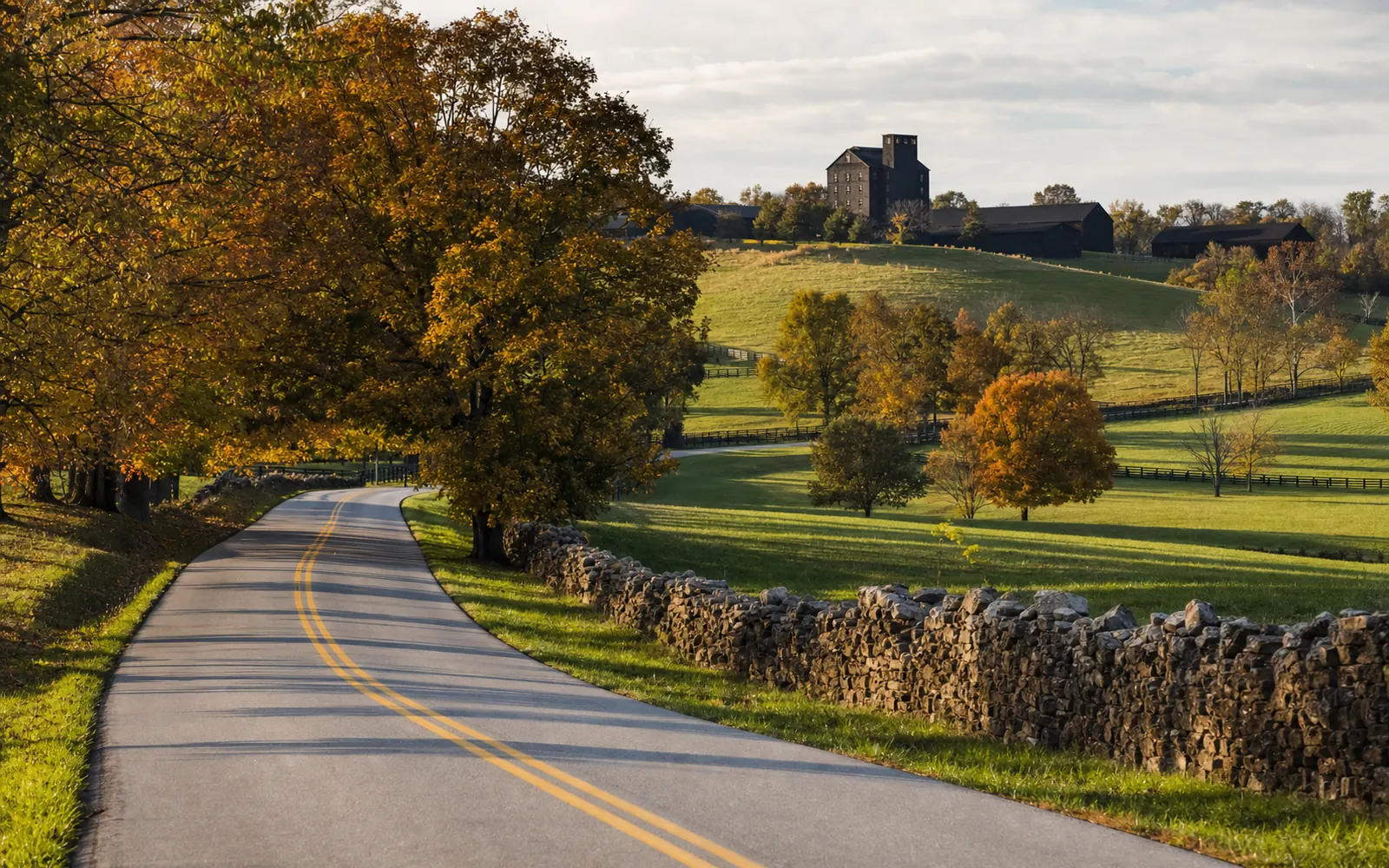 A two-lane country road curving between stone fences and rolling fall pastures toward a distant dark rickhouse, AI-generated illustration