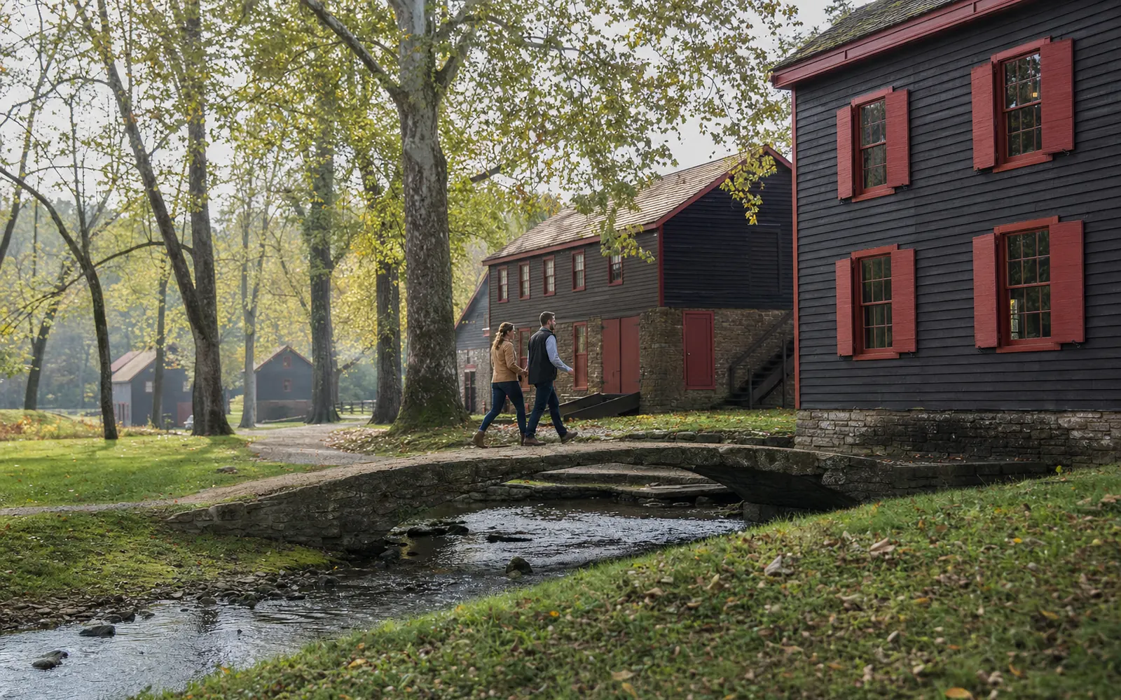 Dark-brown clapboard distillery buildings with red shutters beside a small creek and stone bridge on a green autumn morning, AI-generated illustration