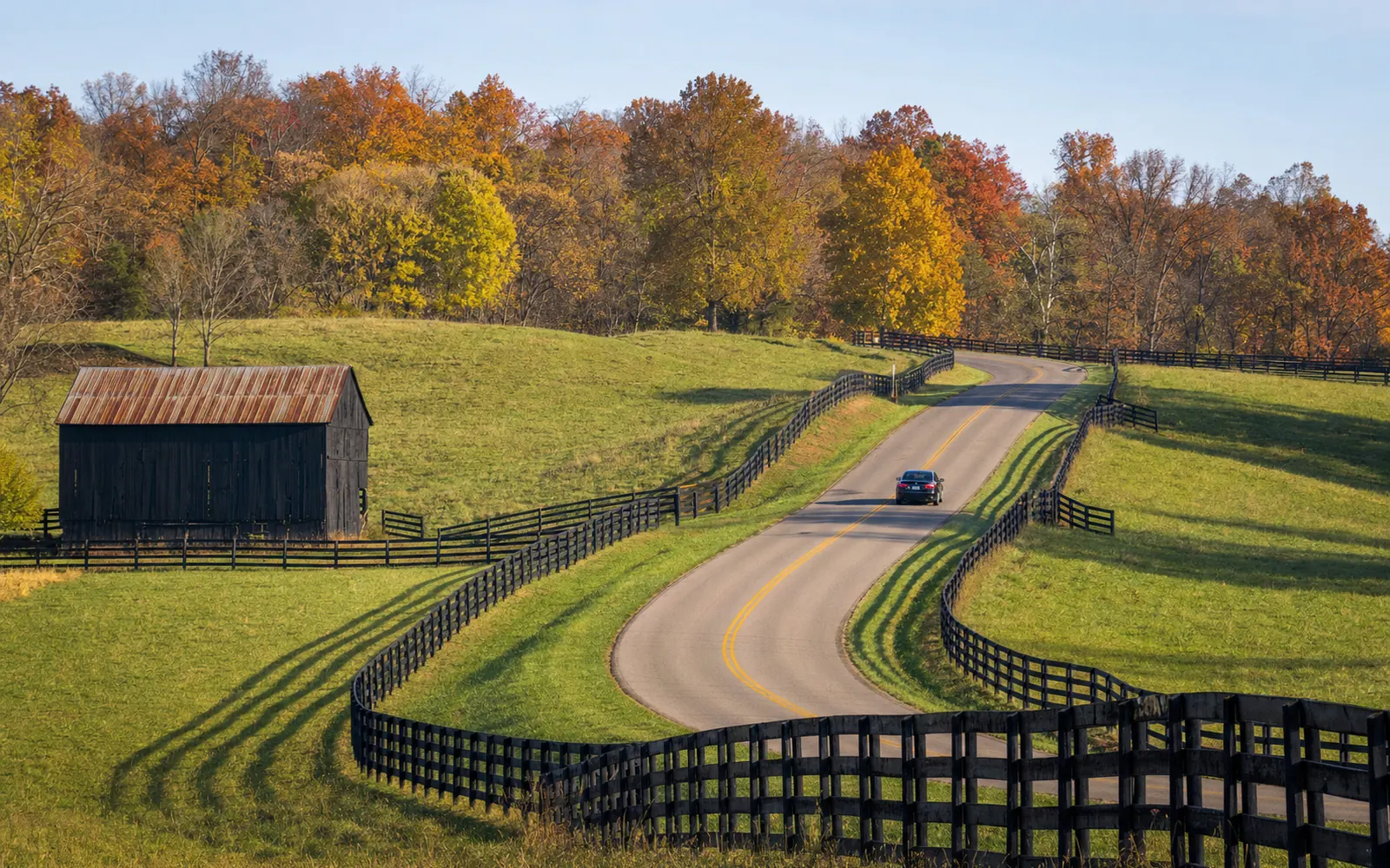 A two-lane country road curving between plank-fenced pastures and autumn woods in central Kentucky, AI-generated illustration