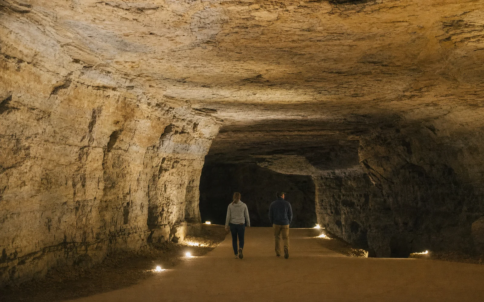 Wide limestone cave passage lit by low electric lights along a paved path, with two visitors in jackets walking into the dark, AI-generated illustration