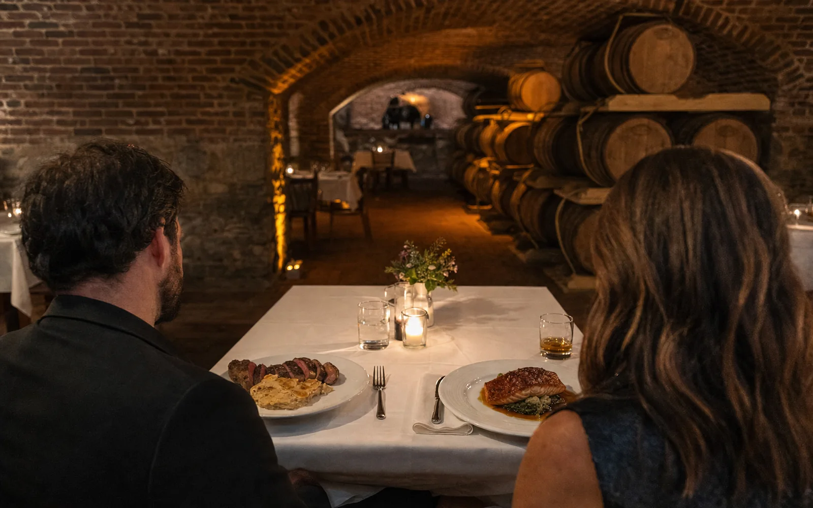 A candlelit table for two in a brick-arched cellar restaurant with steak, salmon and two glasses of bourbon, AI-generated illustration