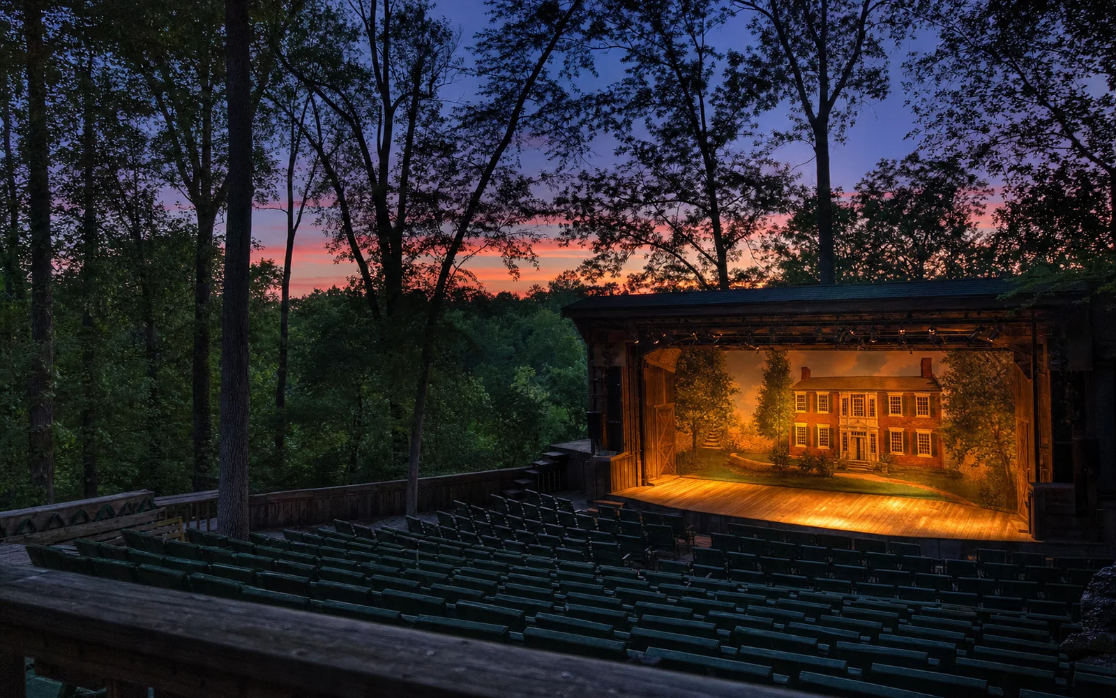 An outdoor amphitheater stage glowing with warm stage lights at dusk, surrounded by tall summer trees, AI-generated illustration