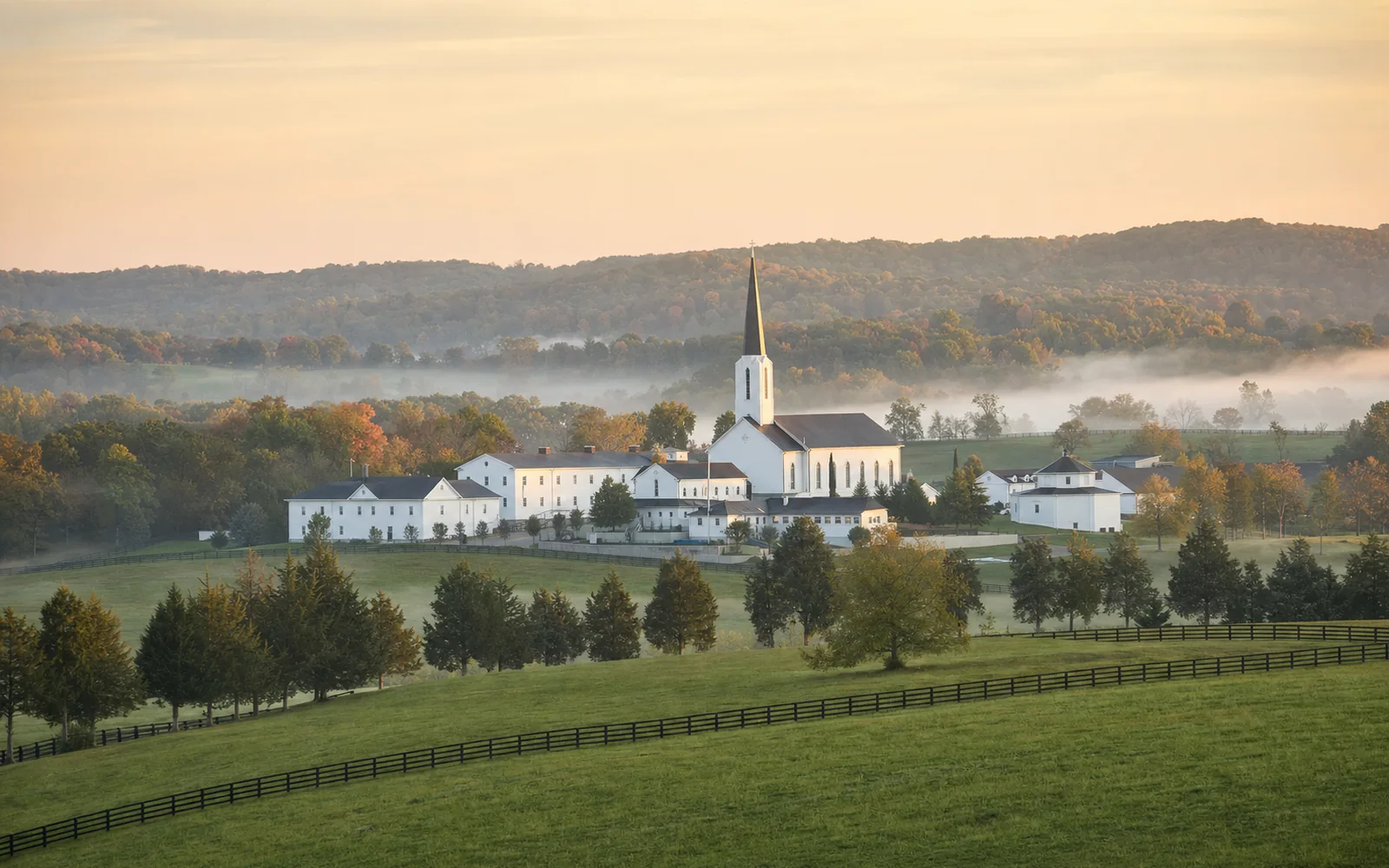 White monastery buildings with a slender church spire in a green valley ringed by wooded knobs, morning mist over the pastures, AI-generated illustration