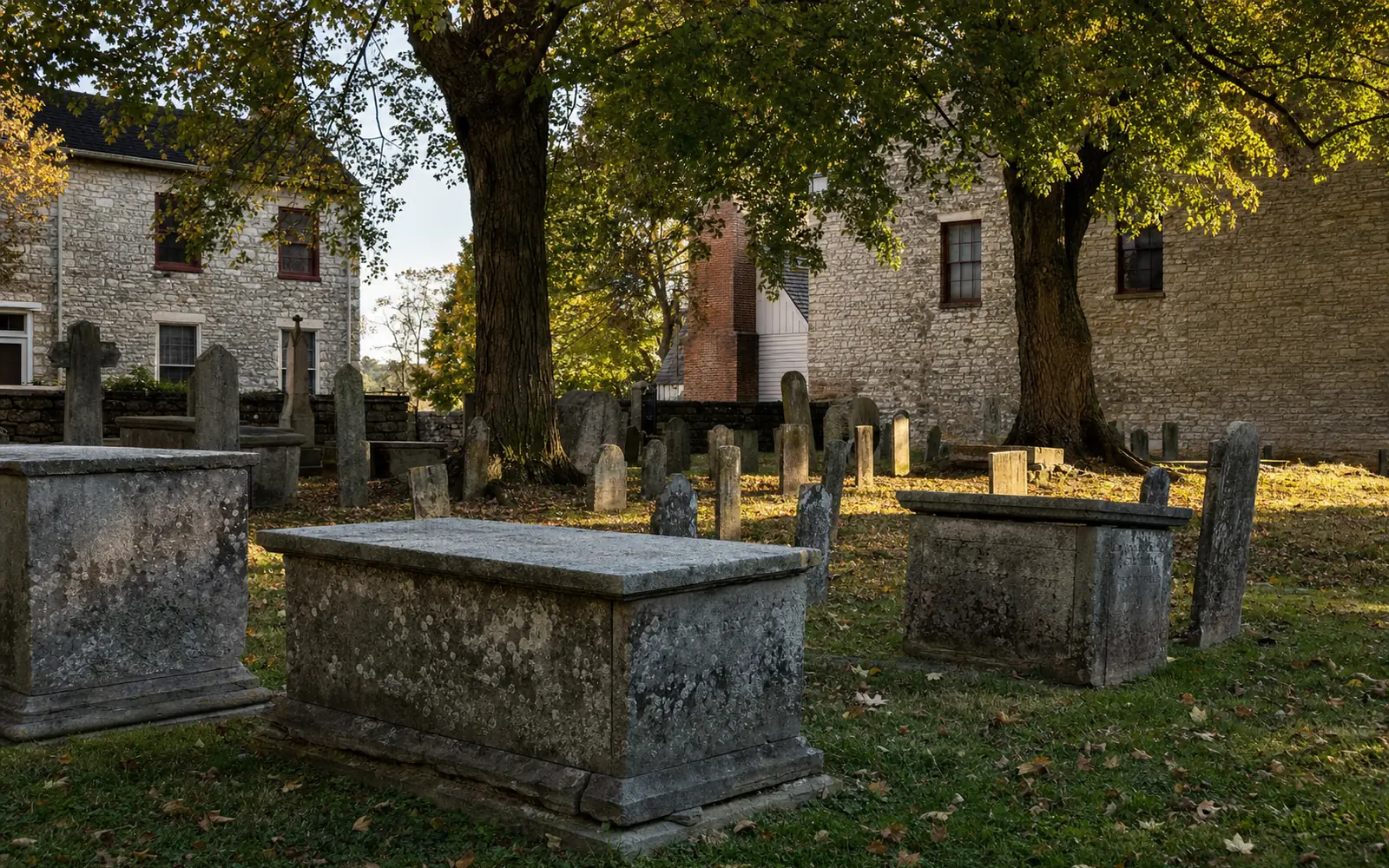 Small old cemetery with weathered limestone chest tombs and leaning headstones under large shade trees beside old stone buildings, AI-generated illustration