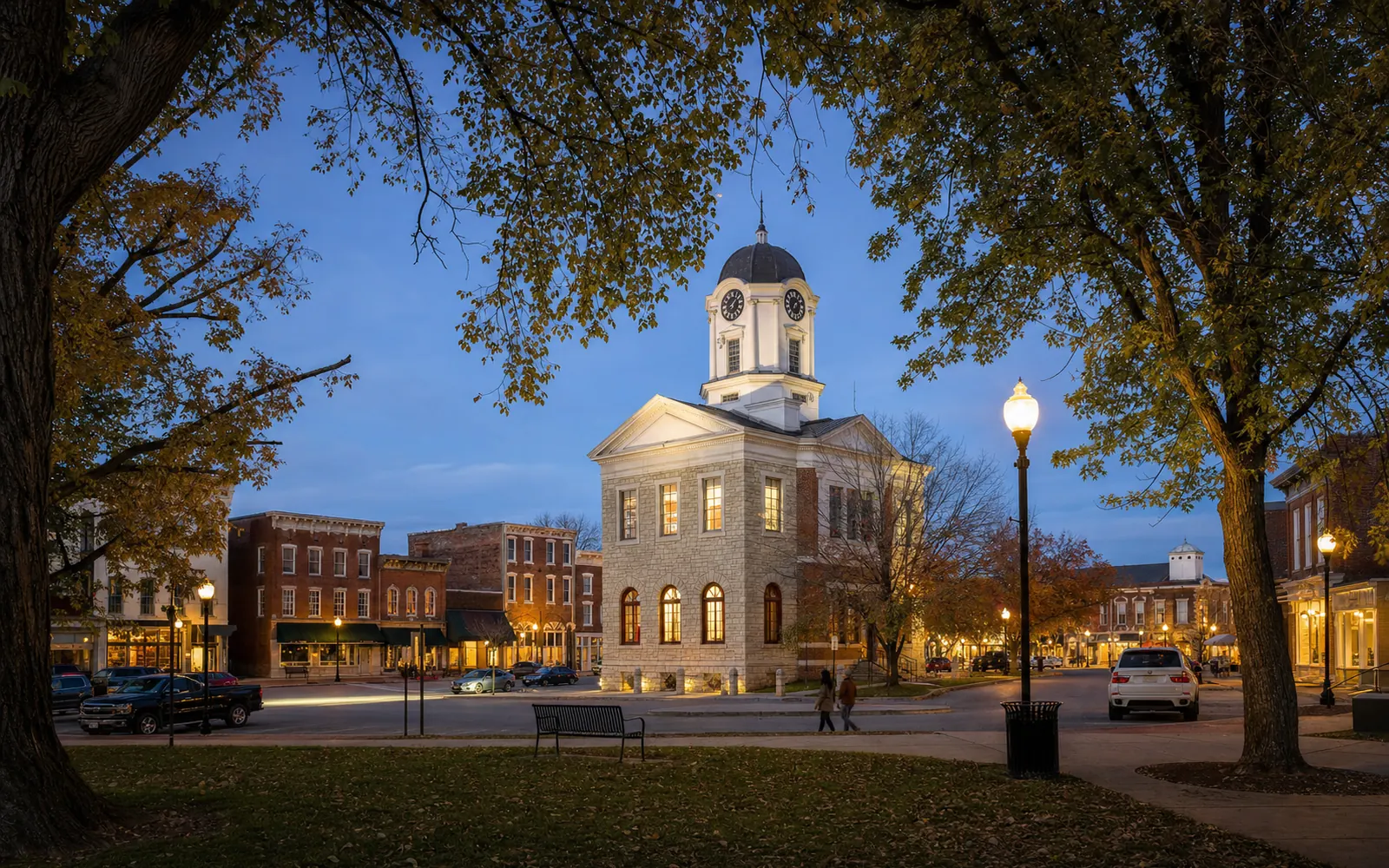 Old stone courthouse with a clock tower in the center of a small-town square at dusk, streetlights just coming on, AI-generated illustration