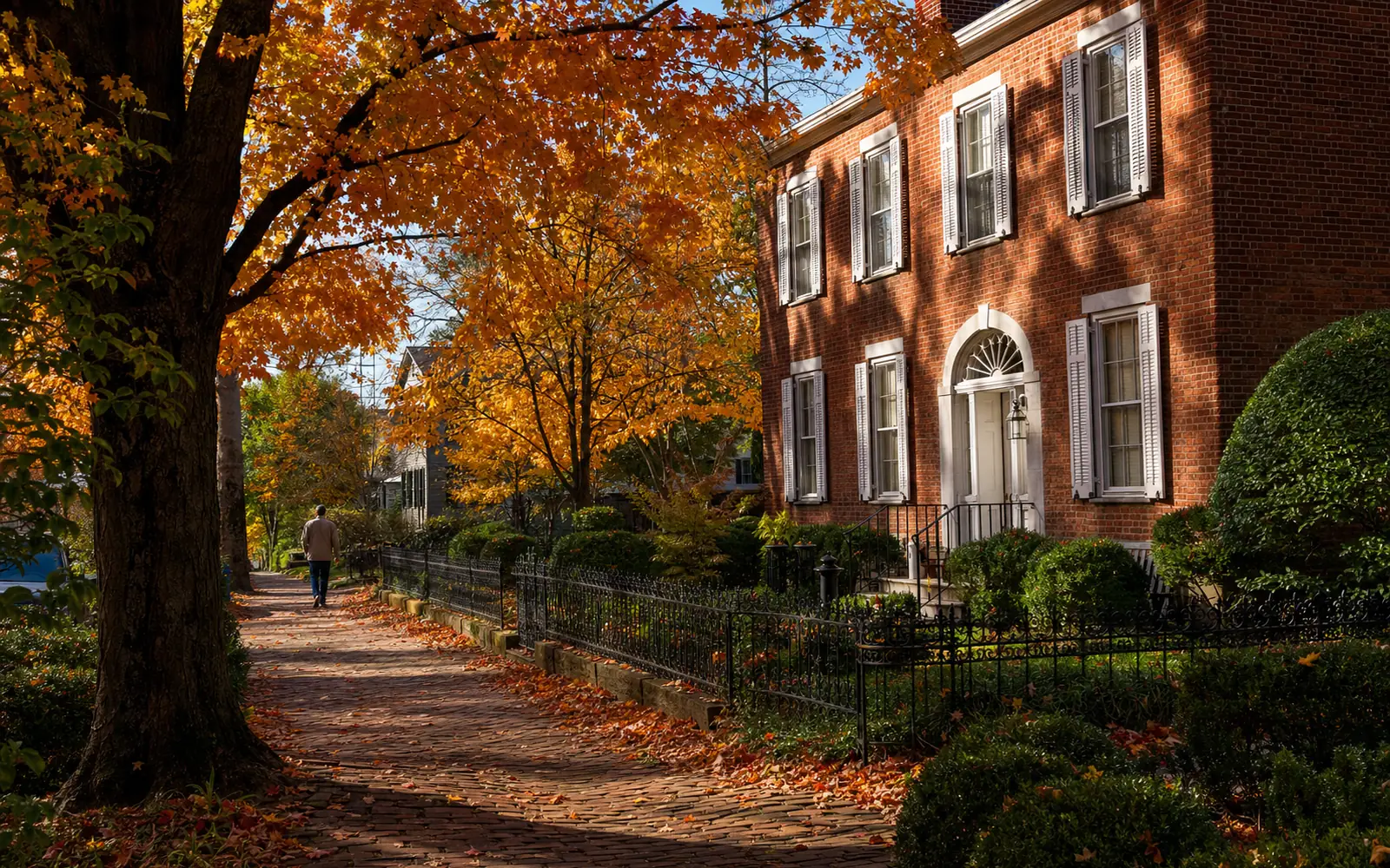Brick sidewalk under orange and red maple trees beside a Federal-style brick house with white shutters on a sunny October afternoon, AI-generated illustration