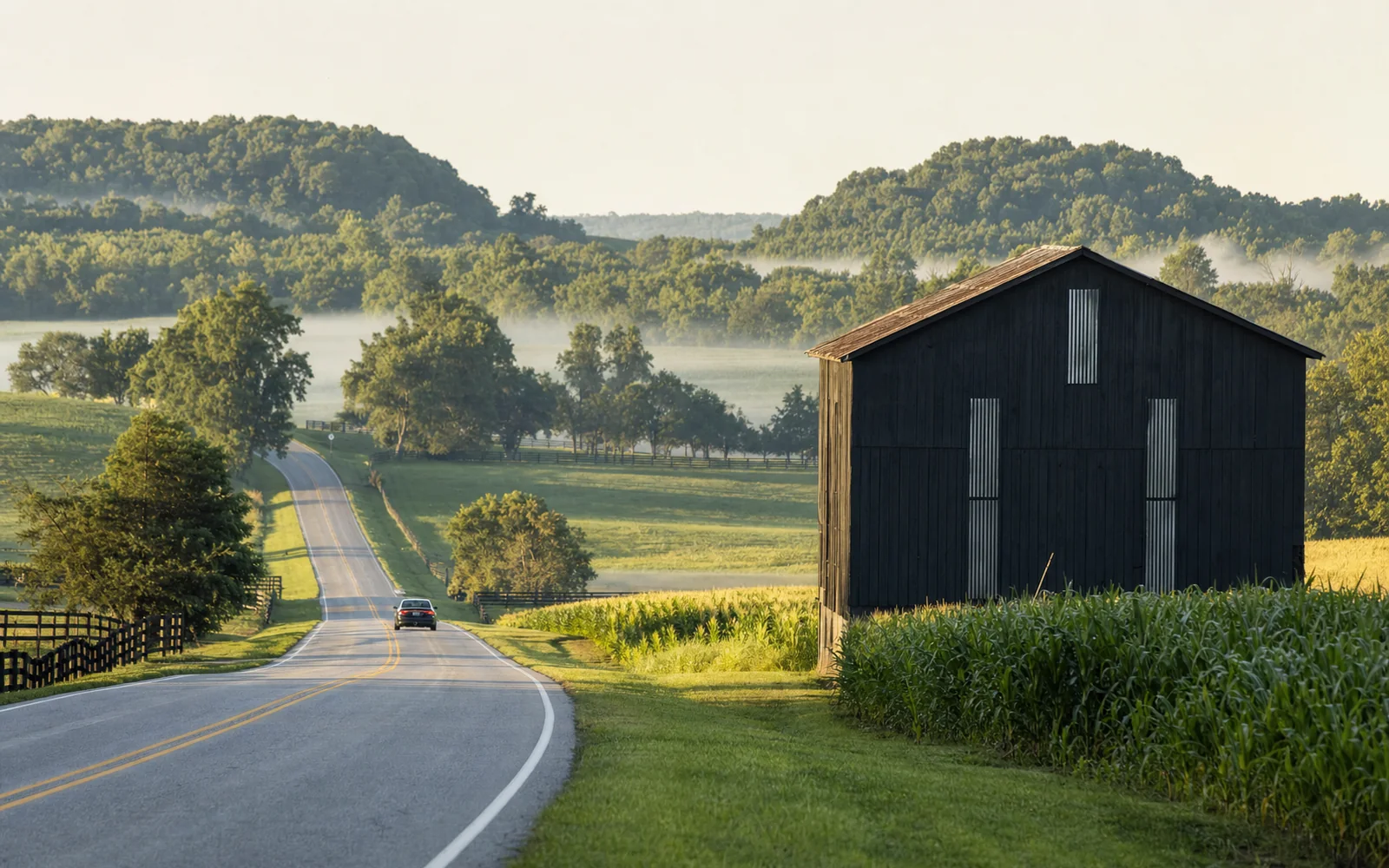 A single car on a two-lane highway through green rolling farmland with a black tobacco barn beside the road in morning light, AI-generated illustration