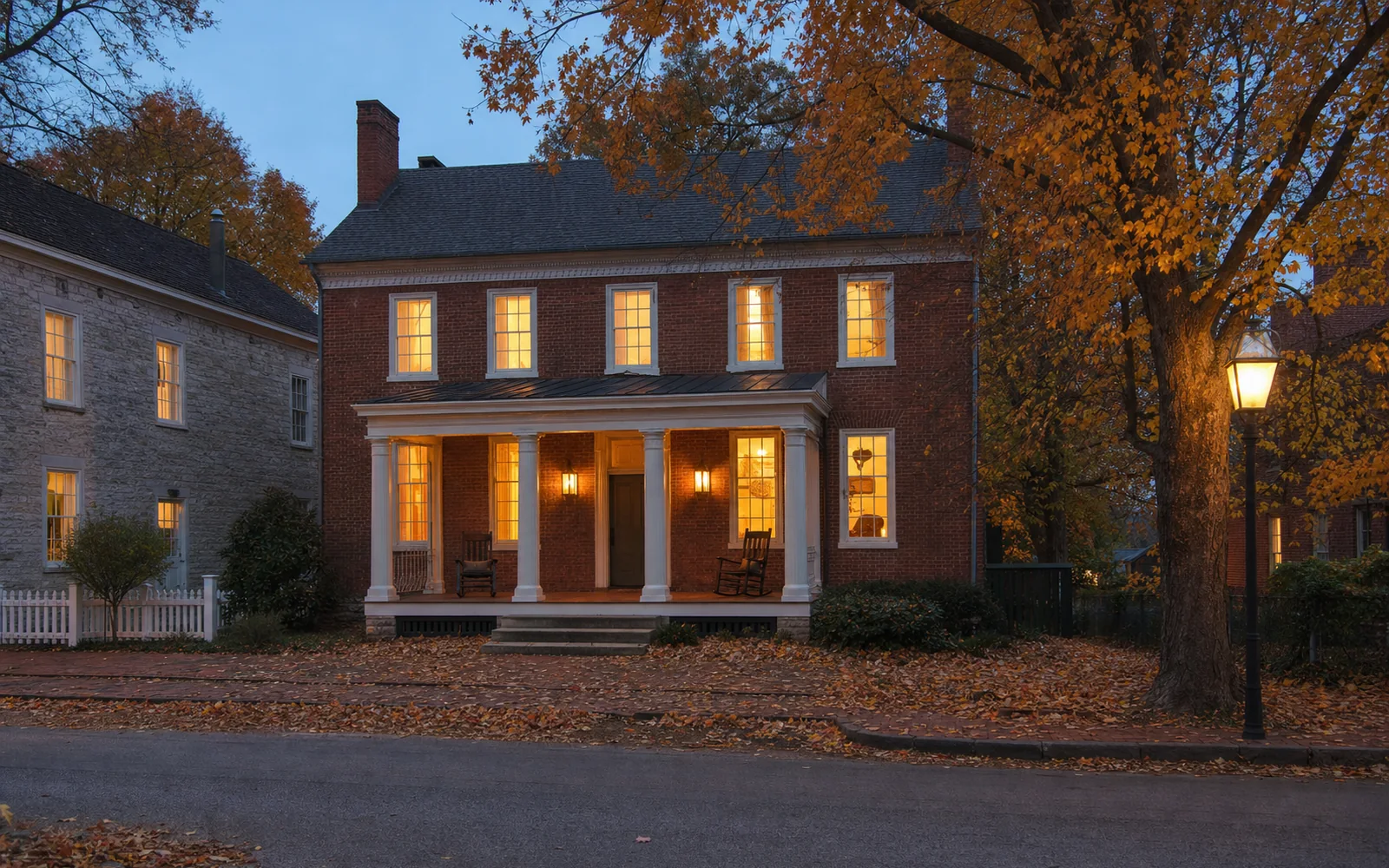 Two-story Federal-style brick house with a white front porch and rocking chairs at dusk, lamplight in the windows, AI-generated illustration