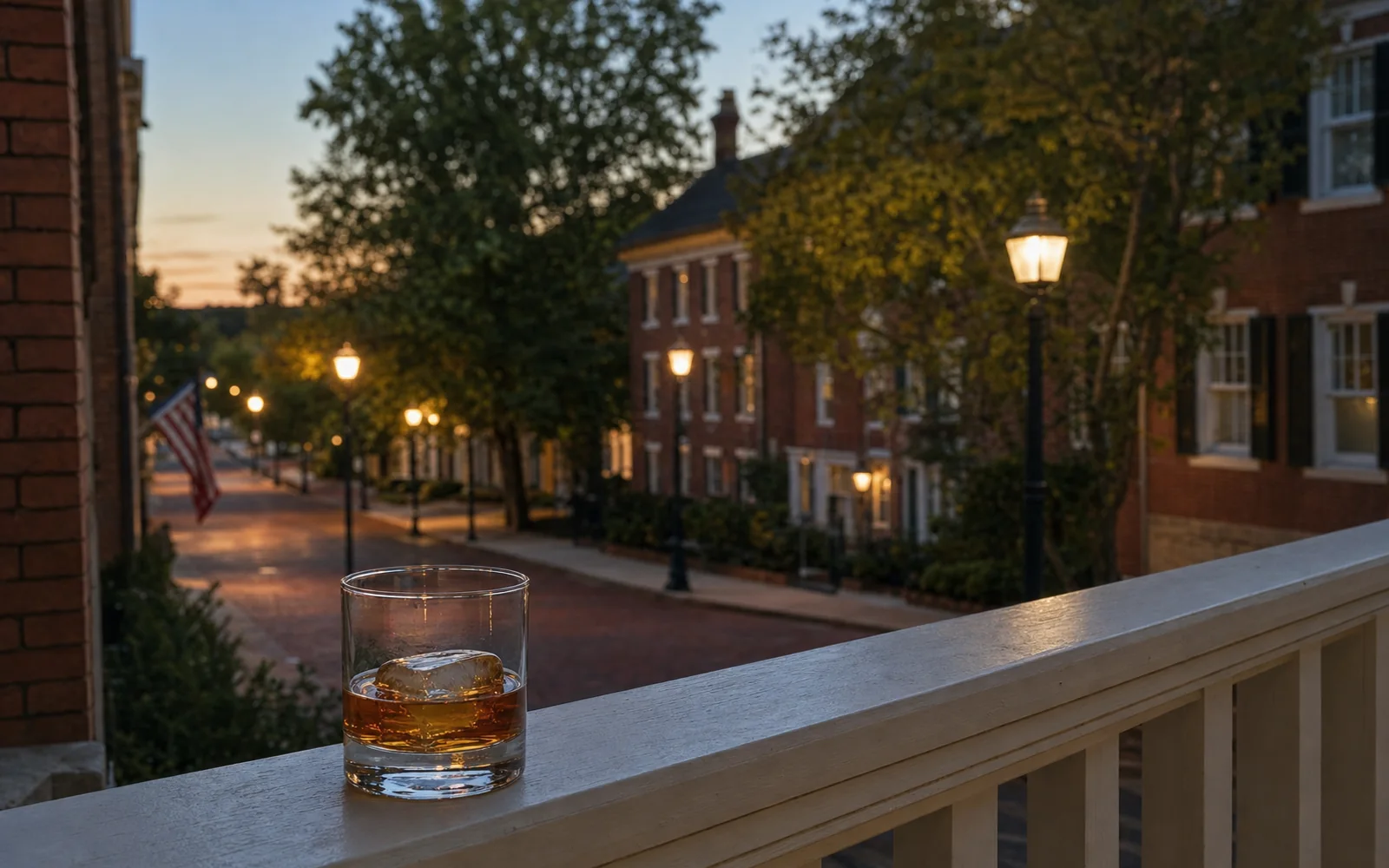 Glass of bourbon on a porch railing of a historic brick inn at dusk, autumn trees and warm street lights beyond, AI-generated illustration