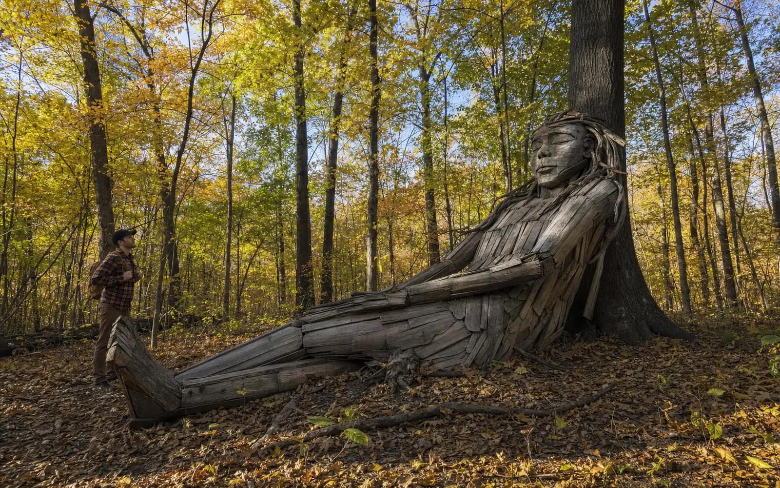 A huge reclining wooden troll sculpture made of weathered reclaimed boards resting against a tree in a sunlit autumn forest, AI-generated illustration