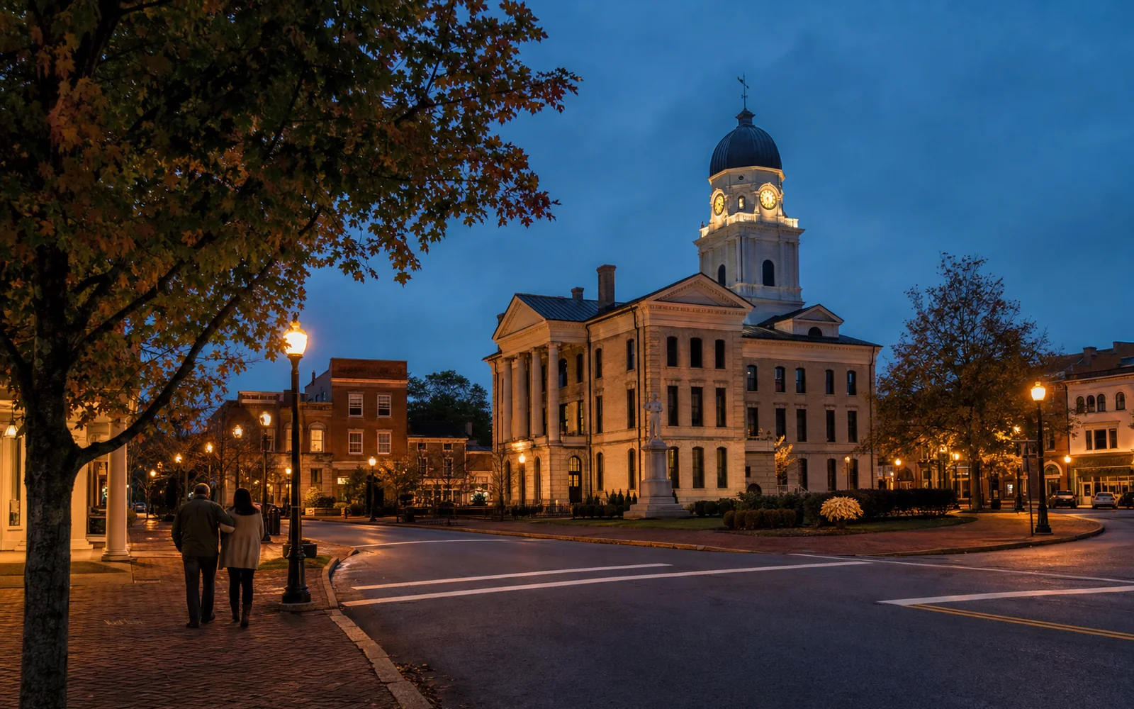 Old courthouse in the middle of a small-town square at dusk, ringed by lit 19th-century brick storefronts with two people strolling, AI-generated illustration
