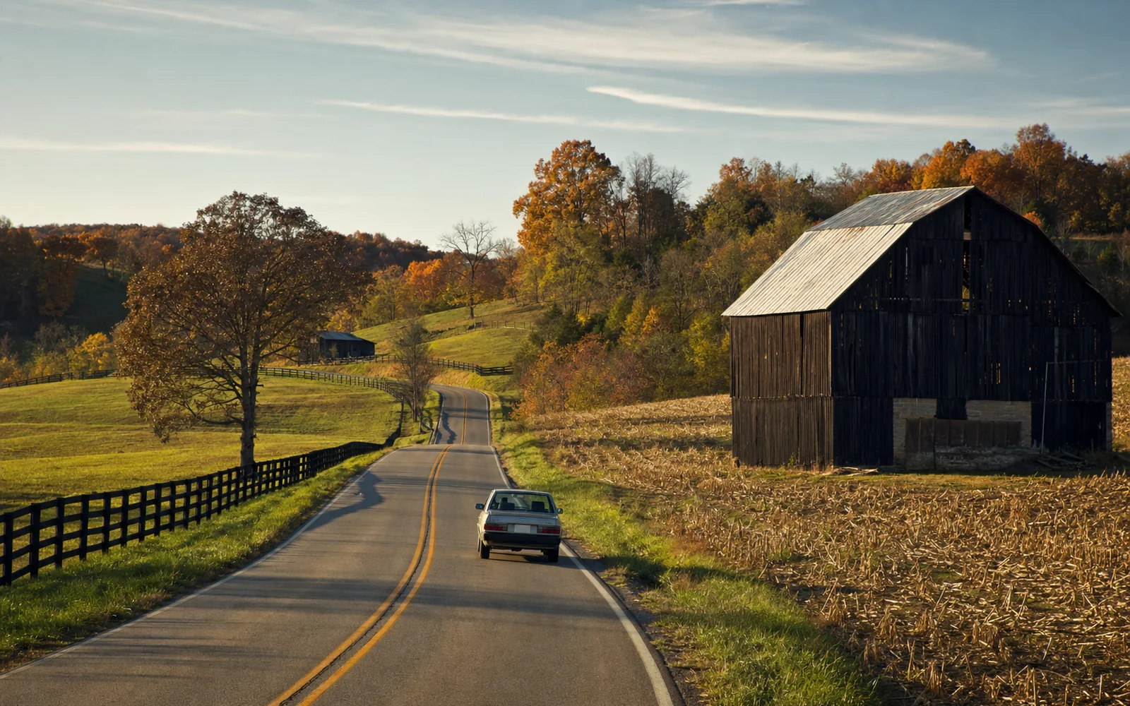 Two-lane country highway curving past a weathered dark tobacco barn and rolling fields with wooded hills in autumn color, AI-generated illustration